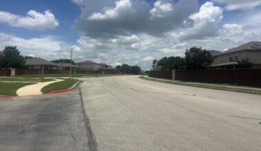 A residential street is shown on a partly cloudy afternoon in Cibolo, TX
