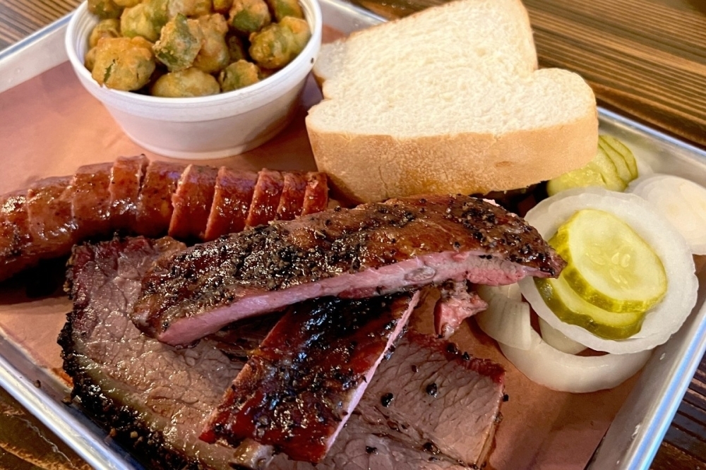 A plate of barbeque meats and bread with a side of pickles and okra