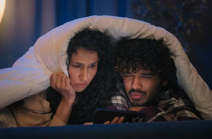 Two people under a blanket look at a phone with puzzled expressions, possibly watching something surprising or intense