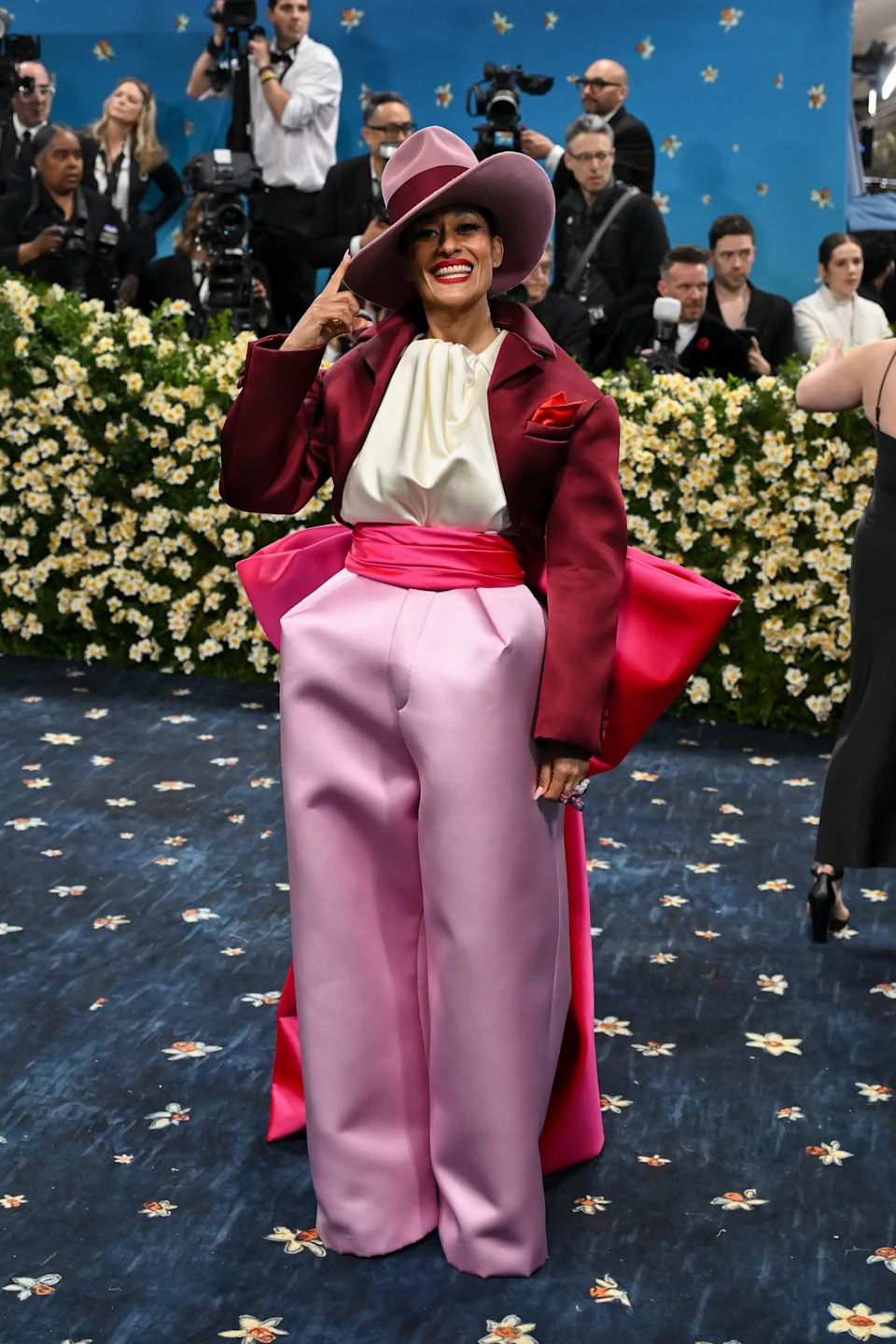 Tracee Ellis Ross in a bold, stylish ensemble with a wide-brim hat poses confidently on a red carpet, surrounded by photographers