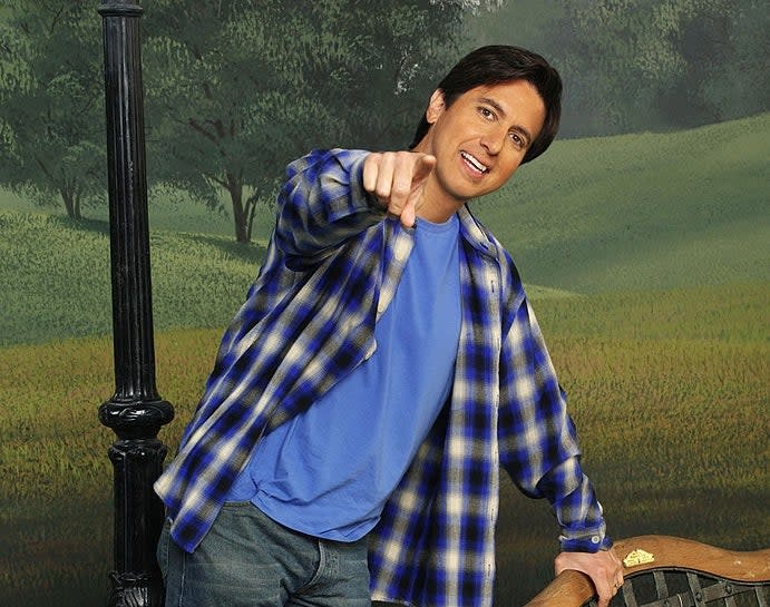 Person in a casual plaid shirt and jeans, leaning on a park bench, smiling and pointing at the camera