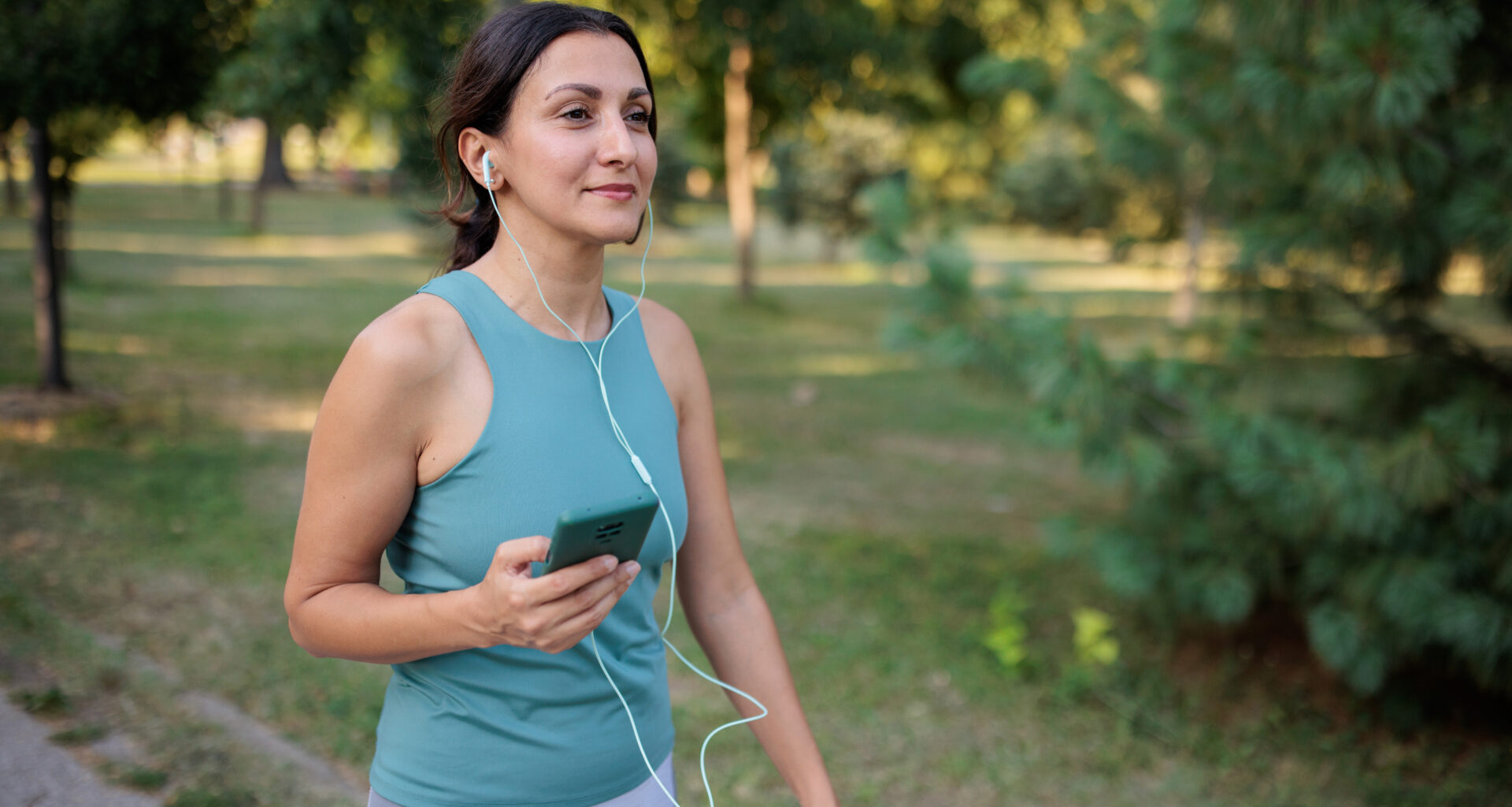 A woman walks outside smiling and wearing headphones, which are plugged into a phone she holds in her right hand. Behind her we see a sunny park with trees.