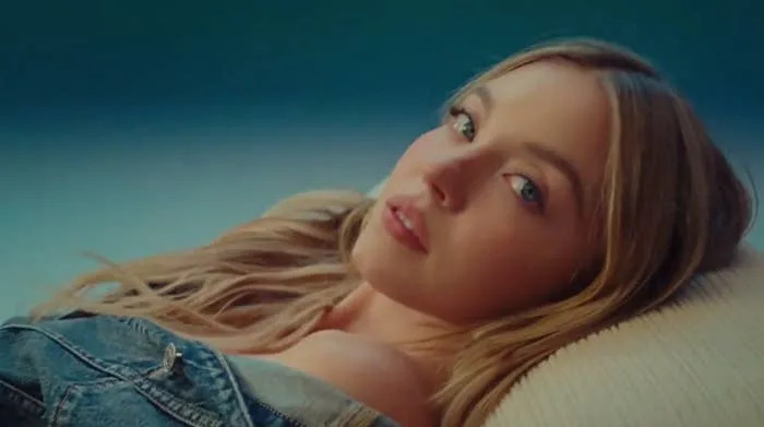 A person with long hair lies on their side, looking towards the camera, wearing a denim jacket off the shoulders