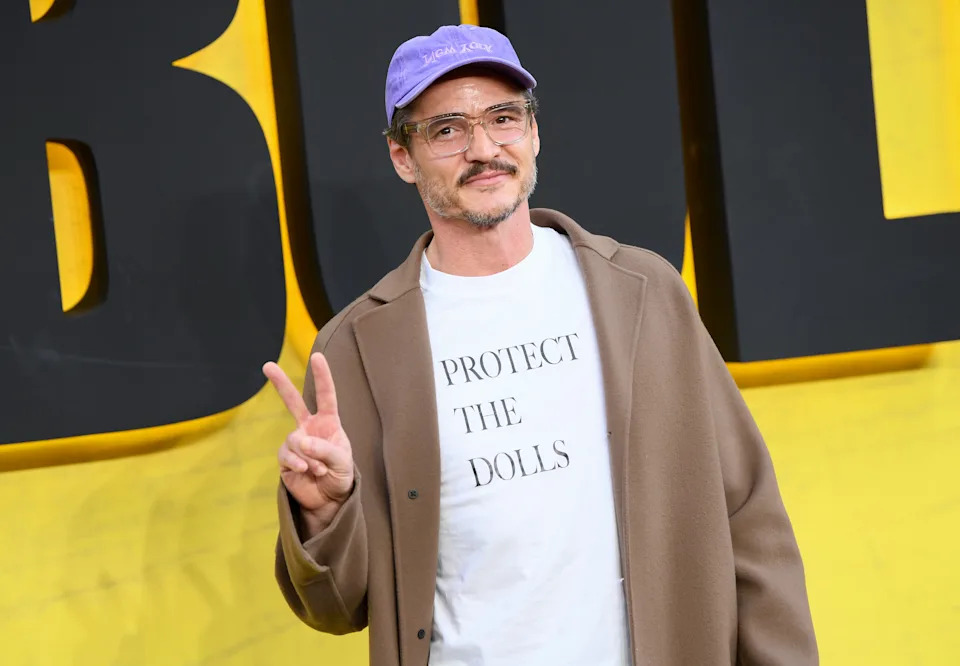 Person on a red carpet in a casual outfit, wearing a cap, glasses, graphic t-shirt, and coat, making a peace sign gesture
