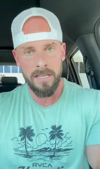 Person with a beard and a cap is sitting in a car, wearing a T-shirt with palm trees and sunset graphic