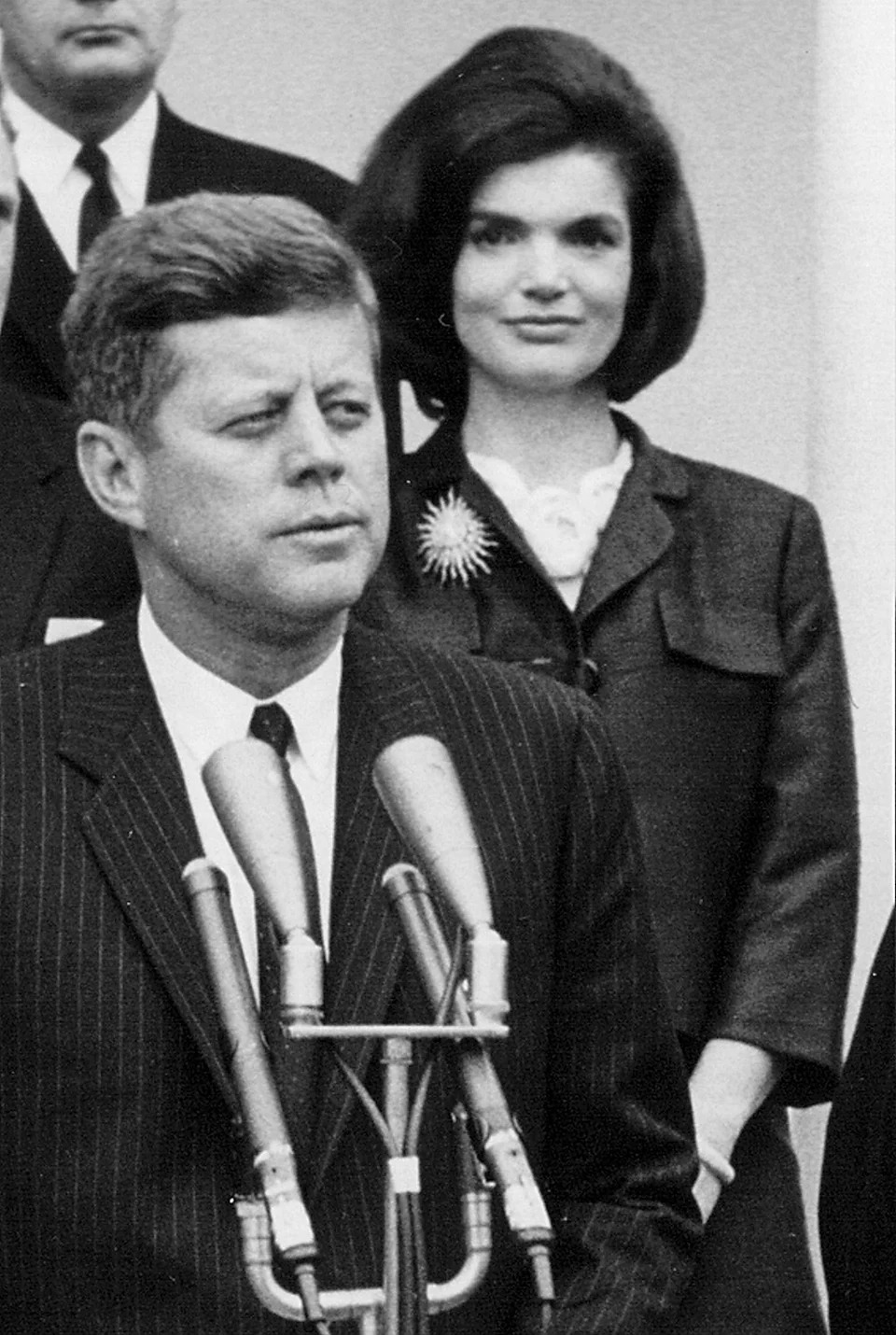 John F. Kennedy speaking at a podium as Jackie Kennedy watches.