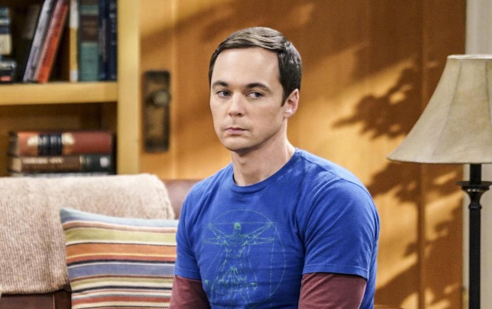 Two characters from a TV show sit on a couch; one in a floral dress, the other in casual attire, surrounded by books and a table of snacks