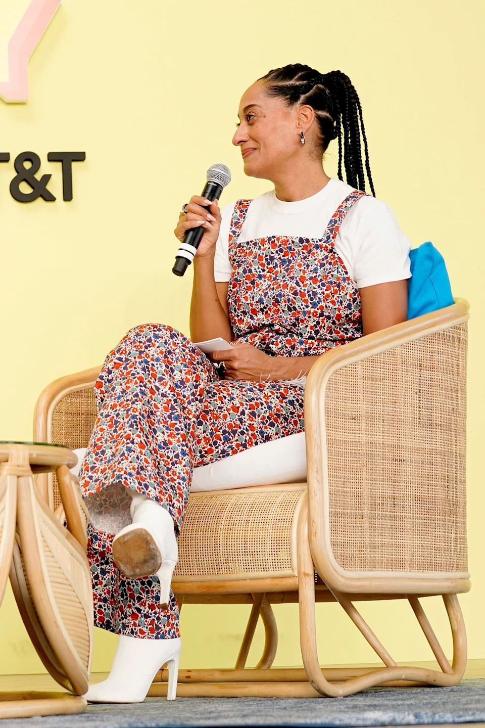 Tracee Ellis Ross seated in a wicker chair, wearing floral overalls, a top, and holding a microphone, engaged in a lively discussion