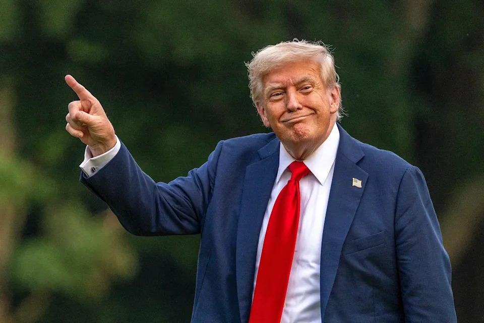 A person in a suit with a red tie gestures with one hand raised and a smile, standing outdoors