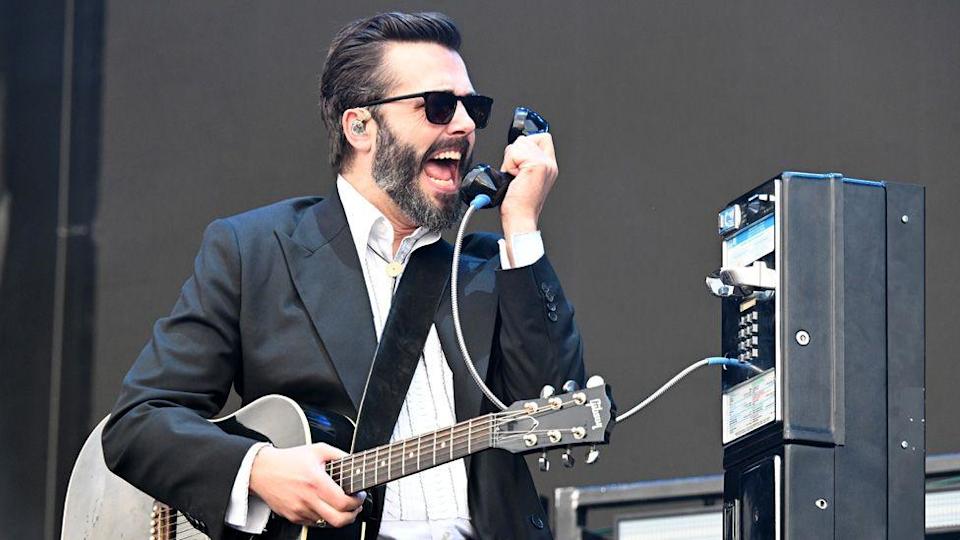 Ben Schneider singing into a payphone handset as a prop on stage, also holding a guitar