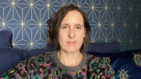 Helen Armstrong A woman with short, dark brown wavy hair is wearing a grey jumper with a neon leopard print pattern. She is sitting on a navy blue sofa in front of a wall with navy and gold-patterned wallpaper.