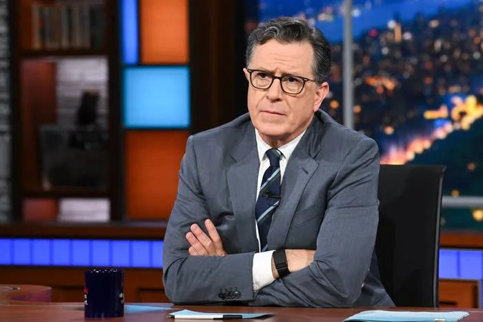A person in a suit sits at a desk on a talk show set, looking contemplative, with arms crossed