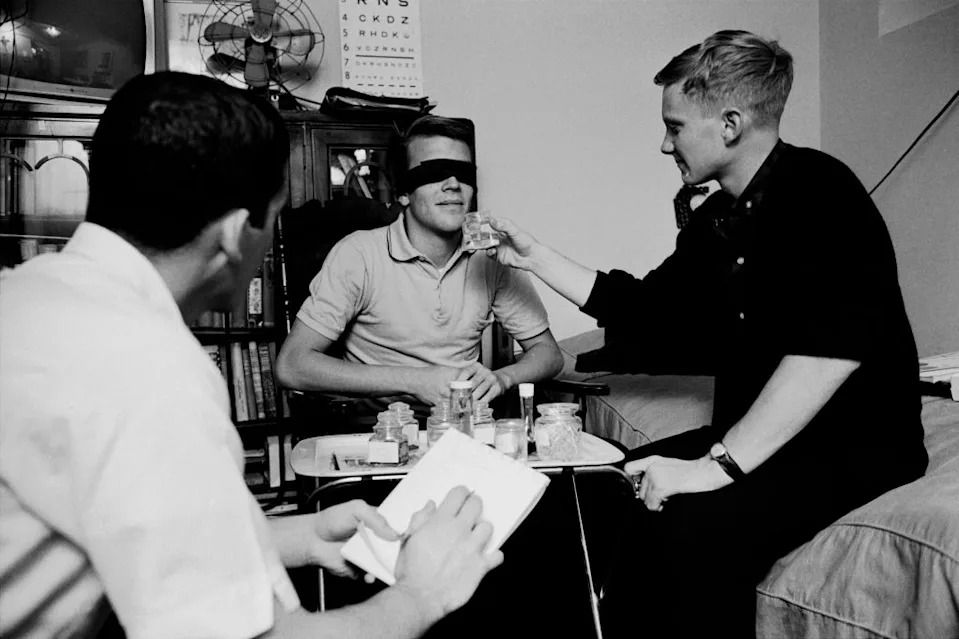 Three men in a room during a scientific experiment; one is blindfolded, another offers him something to smell, while the third takes notes
