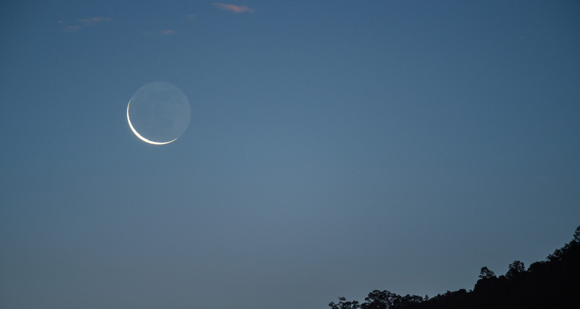 A waning crescent moon is pictured rising in the morning sky above a dark horizon.
