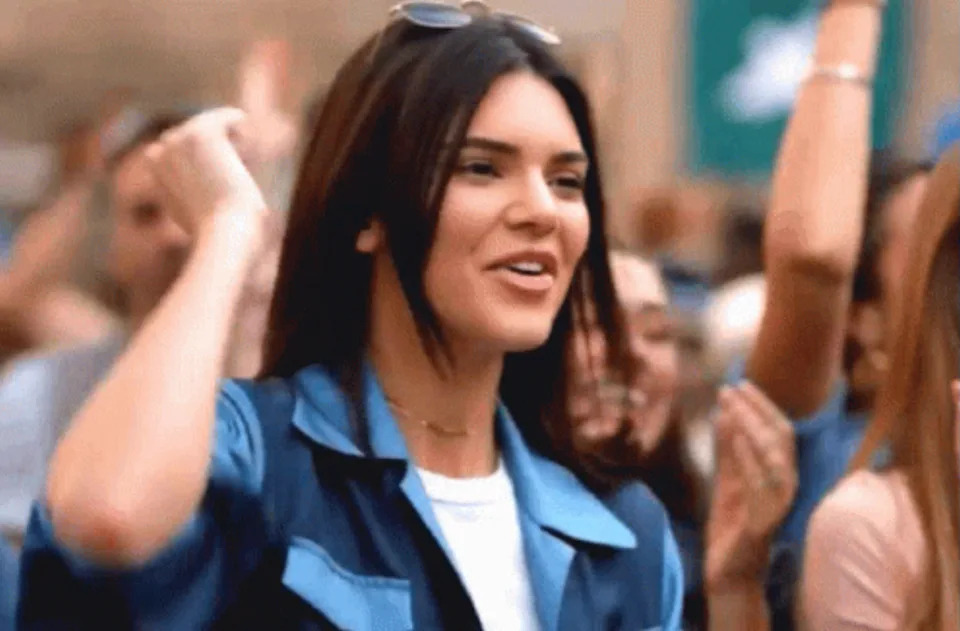 Person energetically cheering outdoors, wearing casual jacket and sunglasses on head, smiling in a crowd