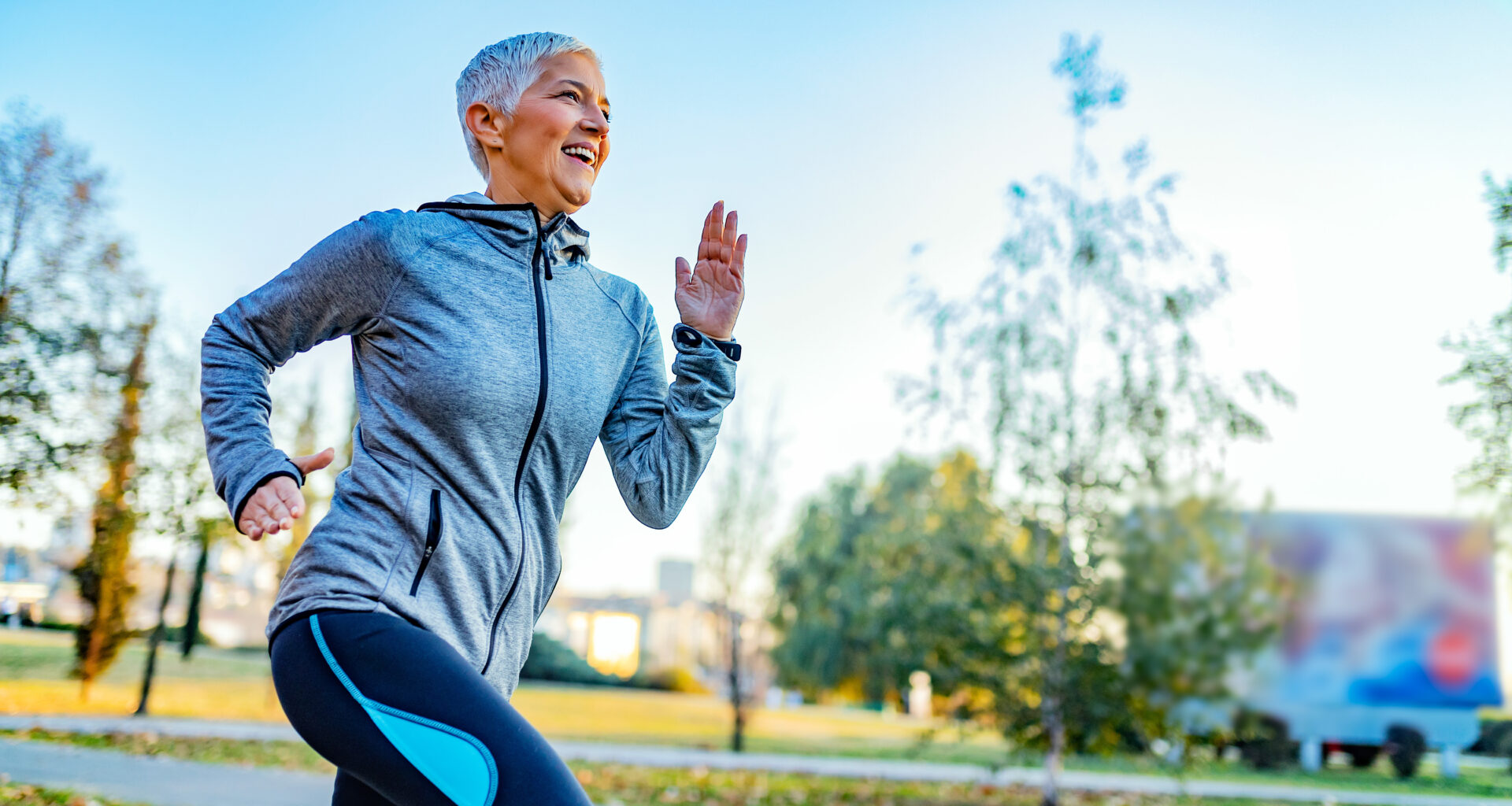 A woman in a sports jacket and leggings smiles as she runs through a park.