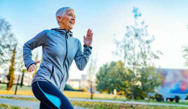 A woman in a sports jacket and leggings smiles as she runs through a park.