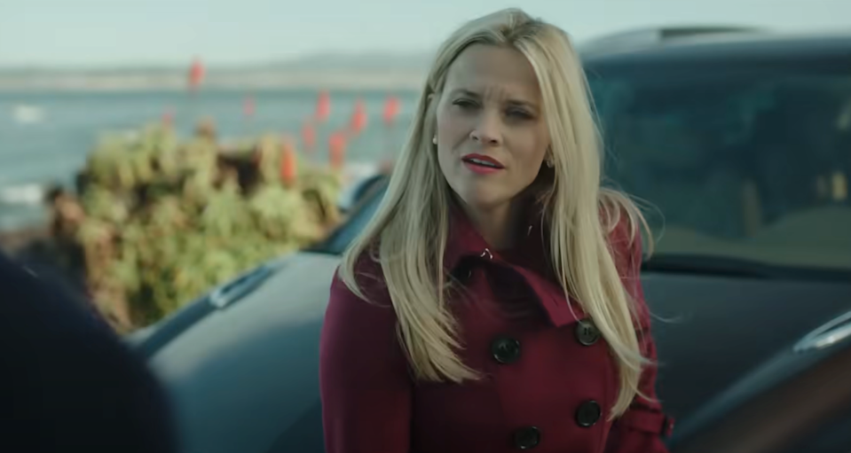 Woman with long hair in a tailored coat stands outdoors near a car, looking engaged