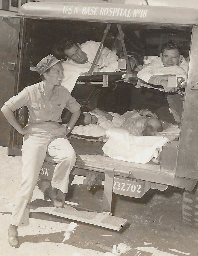 Archival photo of a woman wearing Army fatigues and sitting in the back of a military vehicle with a couple of male military members. The vehicle reads "USN Base Hospital No. 18."