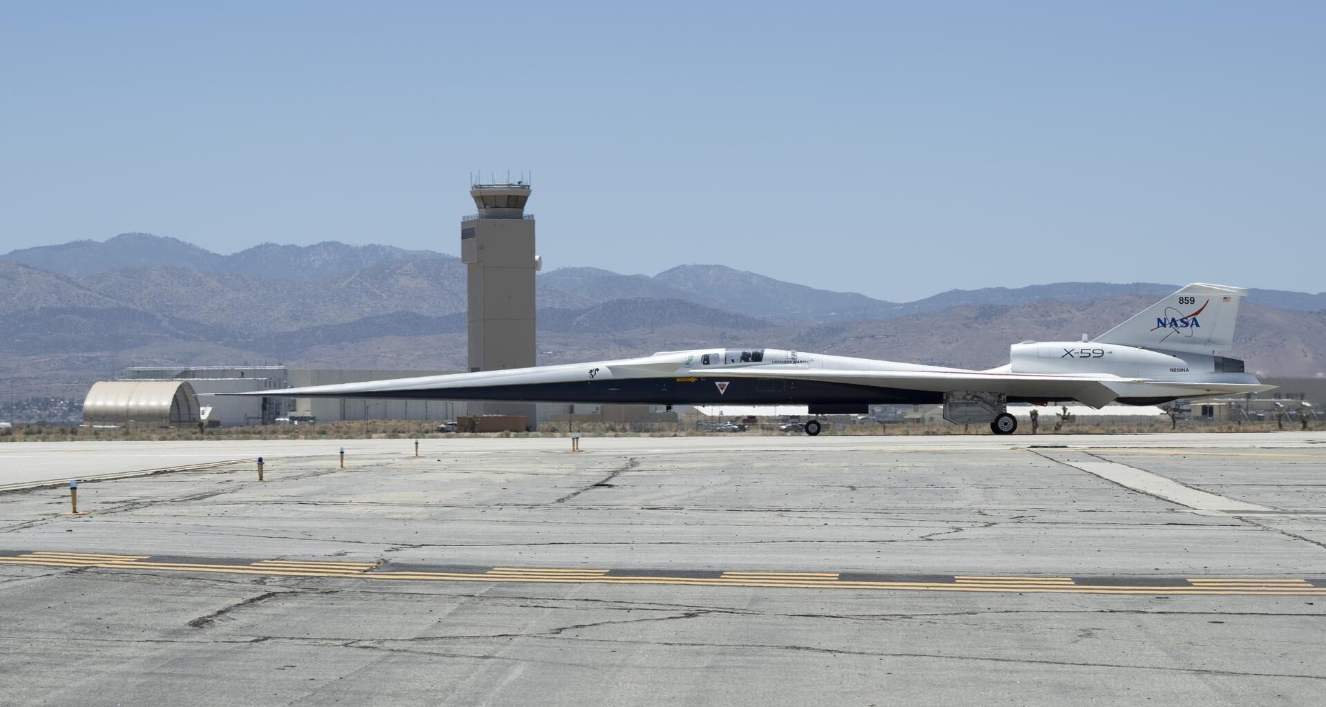 NASA’s X-59 aircraft taxis on the runway at U.S. Air Force Plant 42 in Palmdale, California. The aircraft is shown in profile moving from right to left, with the control tower and desert mountains in the background. In the foreground, the gray concrete runway features a solid yellow line with evenly spaced parallel black stripes inside it, all under a clear blue sky.