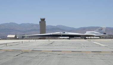 NASA’s X-59 aircraft taxis on the runway at U.S. Air Force Plant 42 in Palmdale, California. The aircraft is shown in profile moving from right to left, with the control tower and desert mountains in the background. In the foreground, the gray concrete runway features a solid yellow line with evenly spaced parallel black stripes inside it, all under a clear blue sky.