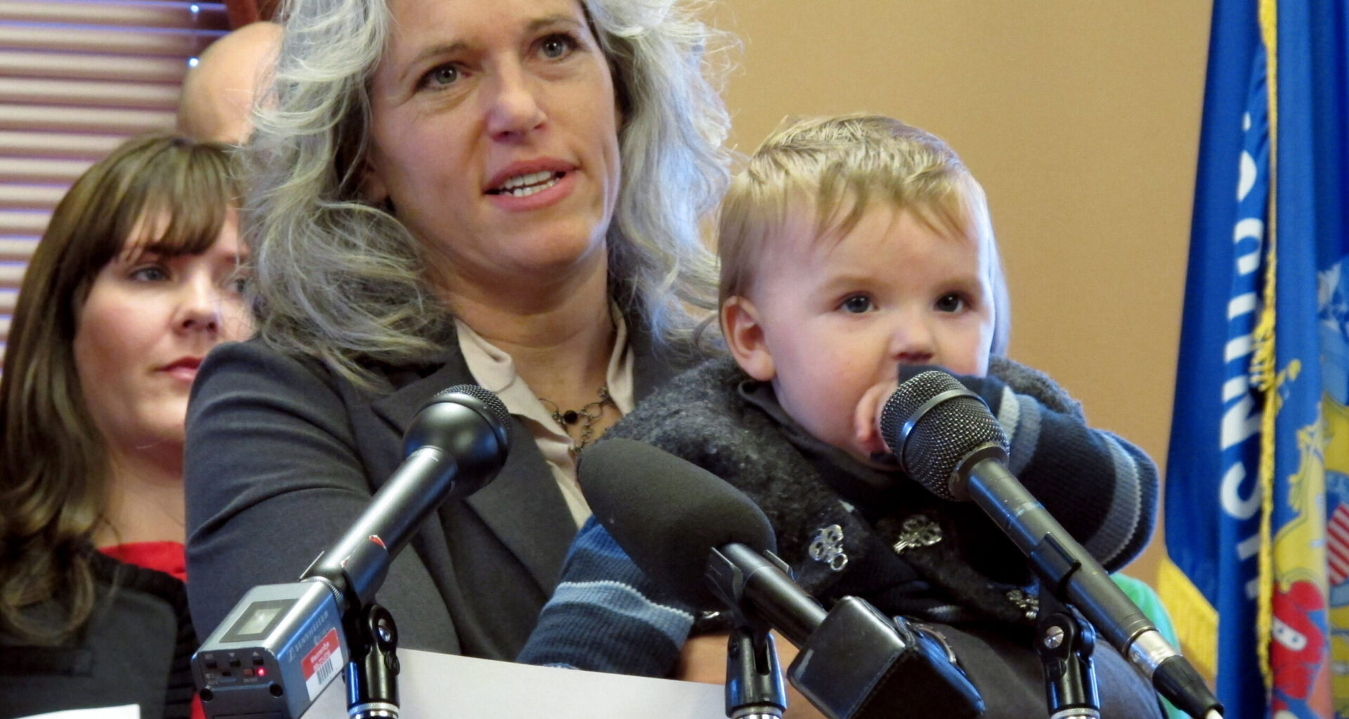 A woman holding a young child stands at a podium with microphones, speaking at an indoor event; a collage of photos is visible on the podium.