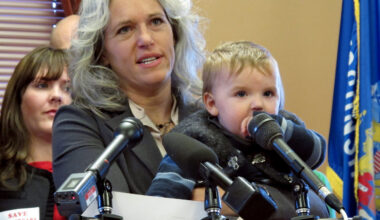A woman holding a young child stands at a podium with microphones, speaking at an indoor event; a collage of photos is visible on the podium.