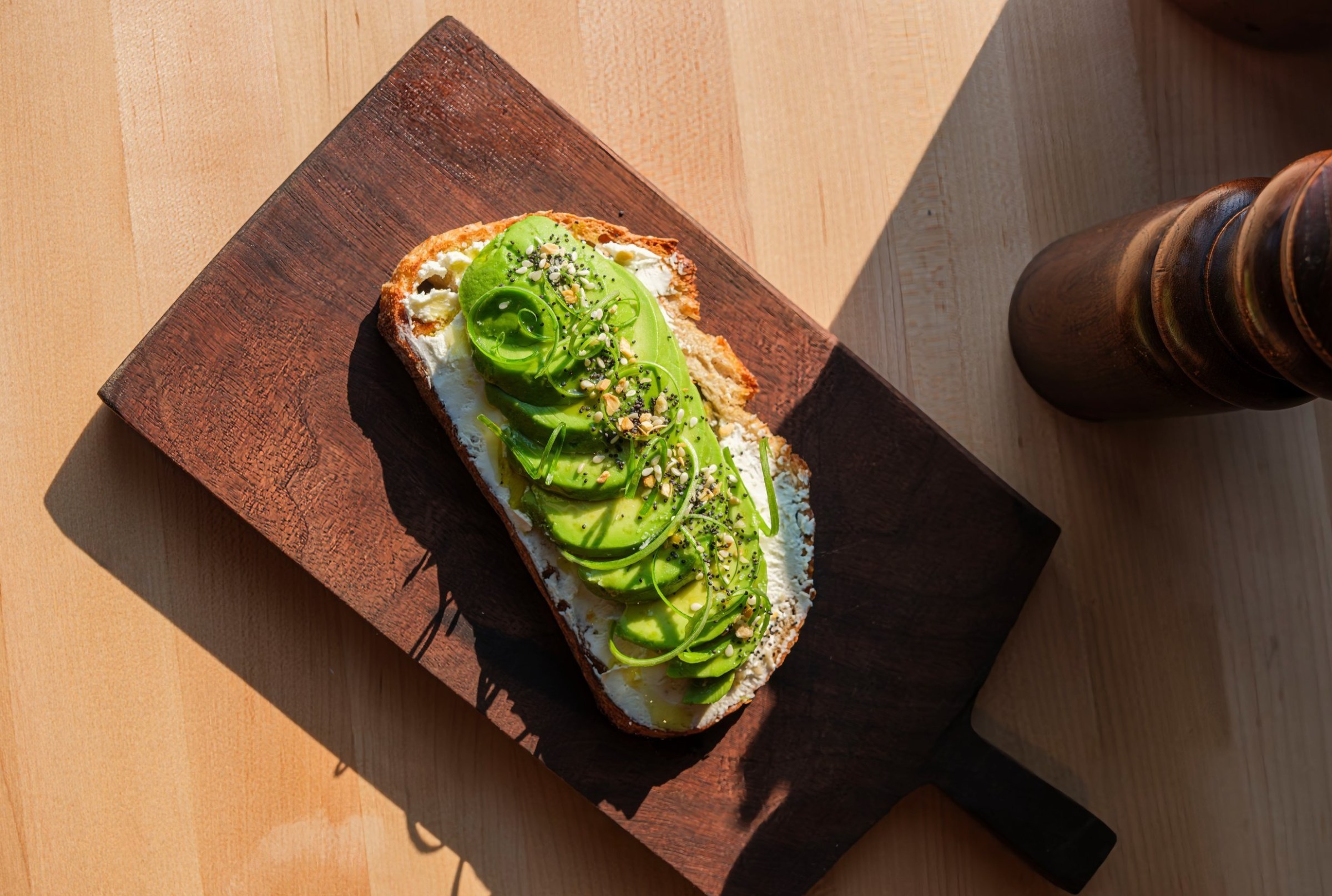 A slide of bread with cream cheese and sliced avocado on a wooden tray on a table.