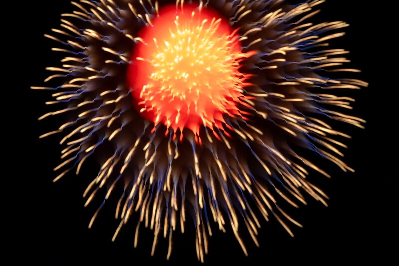 A vibrant firework explodes in the night sky, featuring a bright red center surrounded by radiating yellow and blue streaks against a black background.