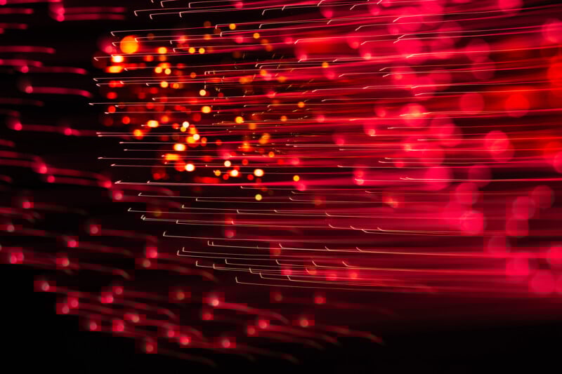 Pink and red streaks and dots of light blur diagonally across a dark background, creating an abstract, vibrant, and energetic pattern reminiscent of fiber optic lights in motion.