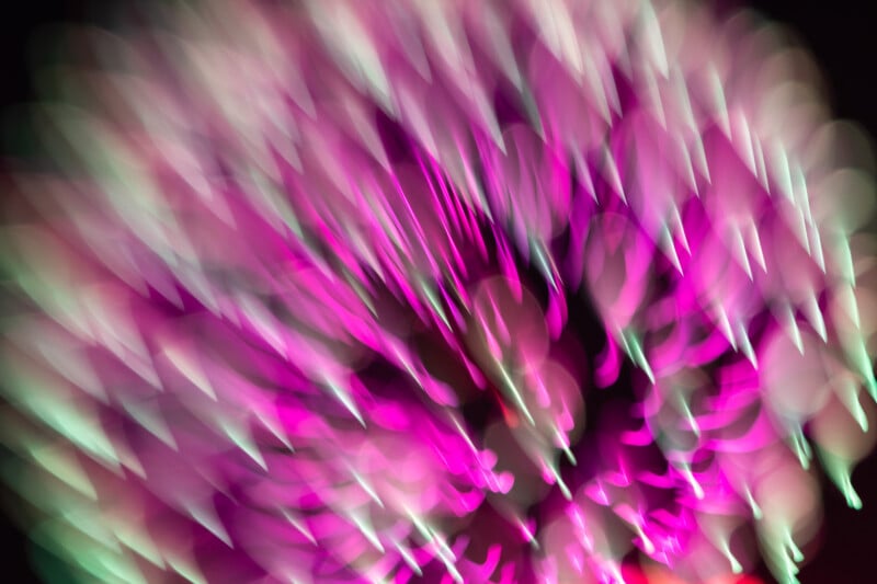 A blurred, abstract image featuring streaks and dots of bright pink, purple, and white light radiating outward, creating a dynamic and vibrant pattern against a dark background.