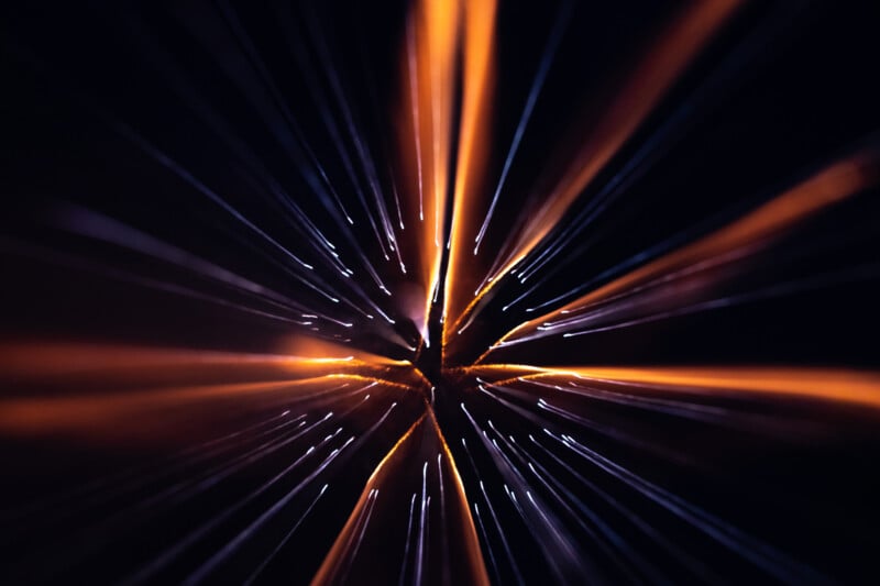 Abstract image showing bright orange and white streaks of light radiating outward from a central point against a dark background, resembling an explosion or starburst effect.