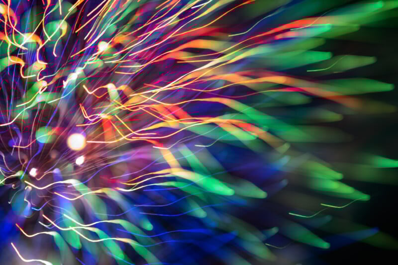 Colorful light trails radiate from a central point on a dark background, creating an abstract, vibrant burst resembling fireworks or energetic motion.
