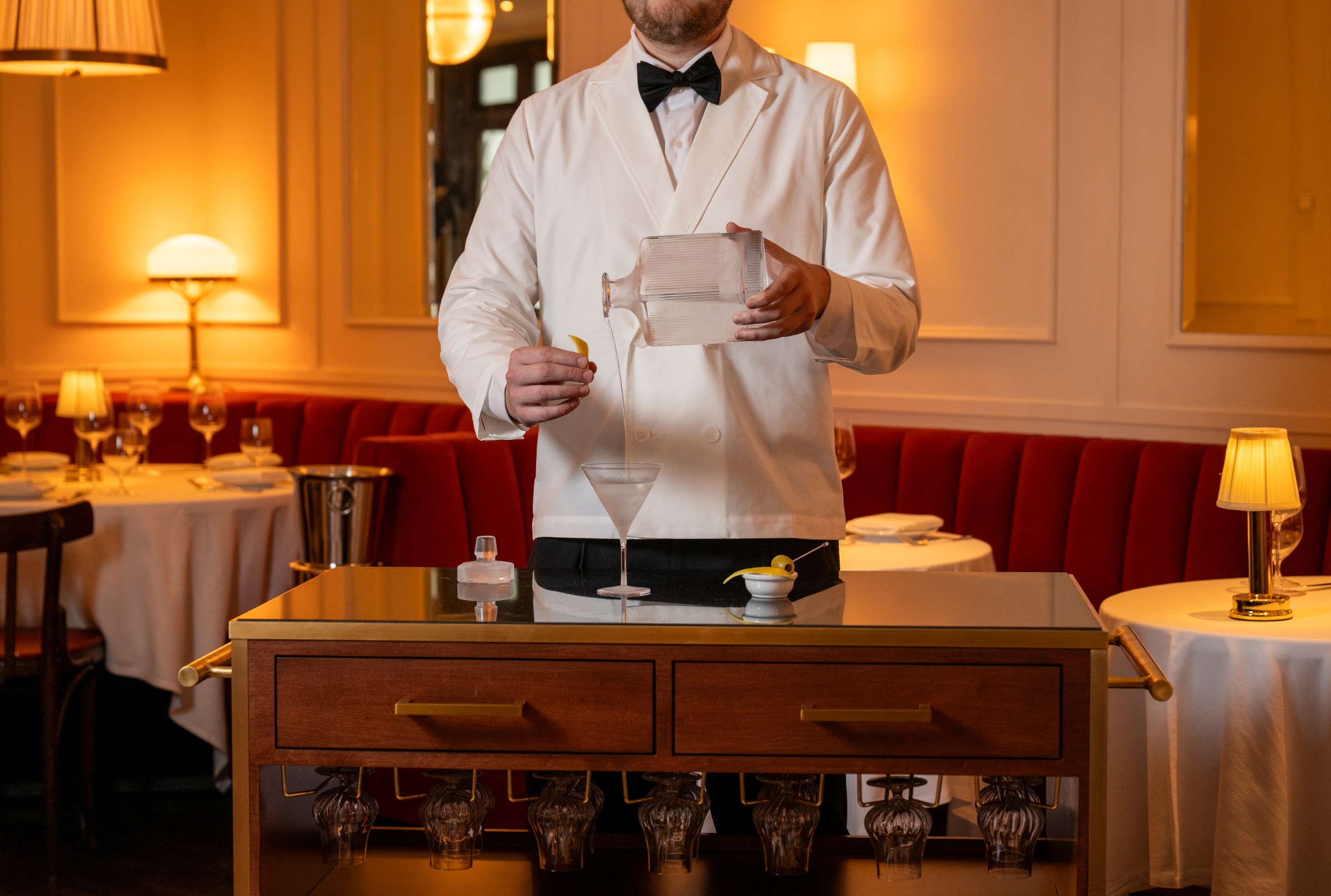 A bartender in a white tuxedo pouring a cocktail into a glass on a cart.