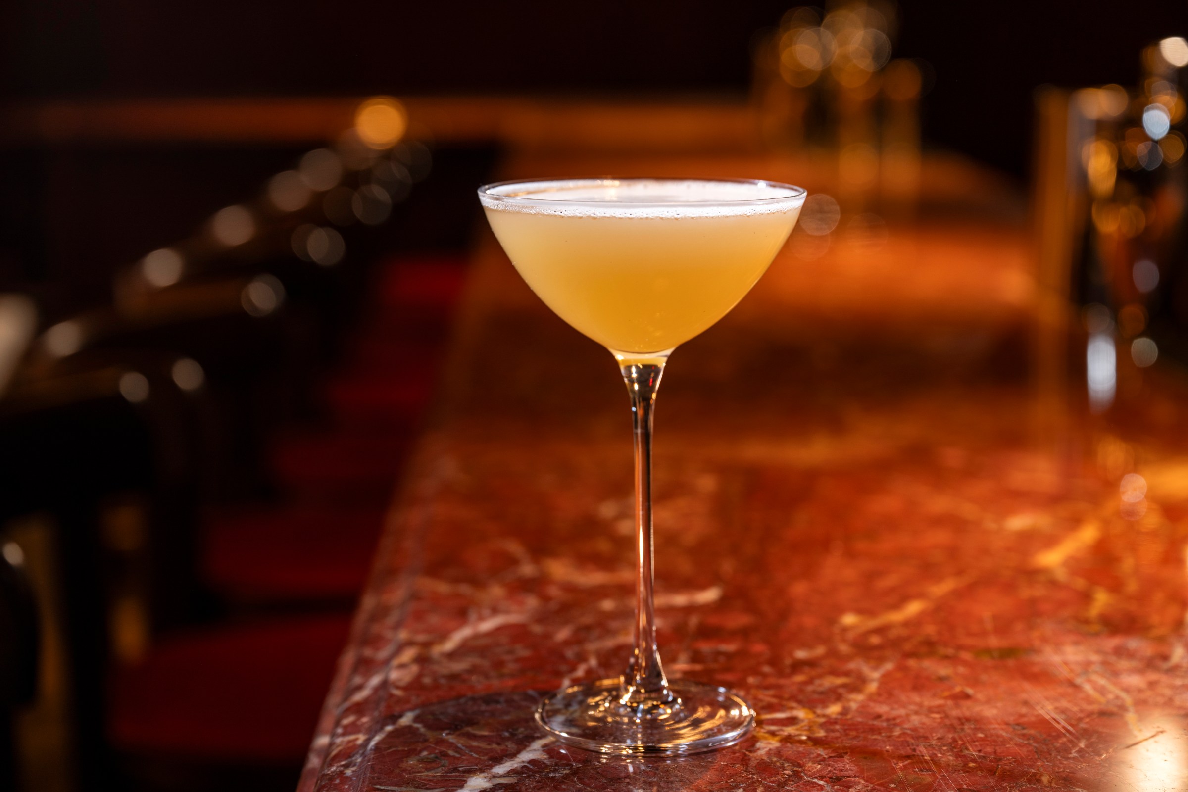 An orange-ish cocktail in a glass on a brown marbled bar.