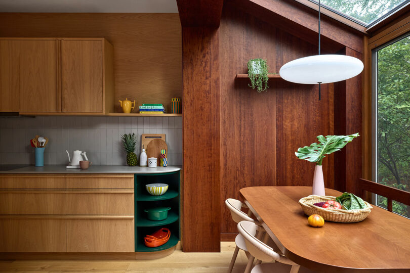 Modern kitchen with wooden cabinets, open shelving, a round dining table with chairs, a skylight, and a basket of fruits and vegetables on the table.