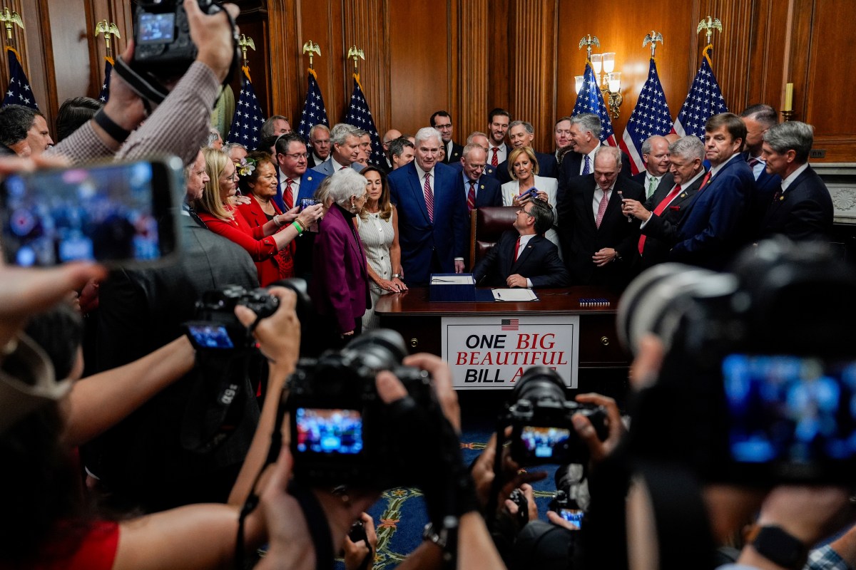A group of officials and photographers surround a person seated at a desk for a bill signing ceremony, with a sign reading "One Big Beautiful Bill" on the front.