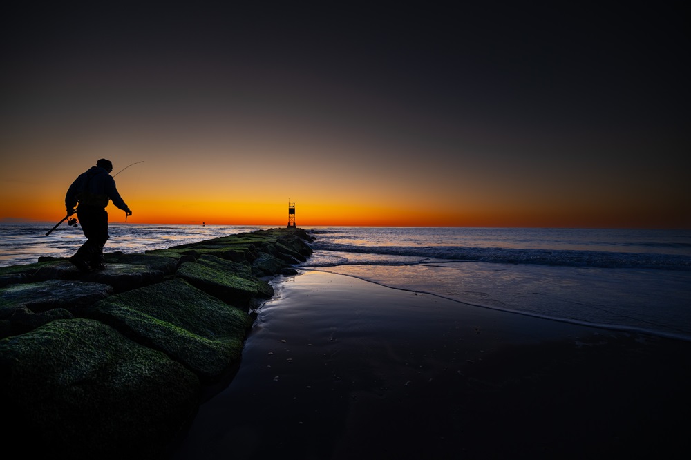A man carrying a fishing rod walks down a breakwater as the sun just beings to peak over the horizon.