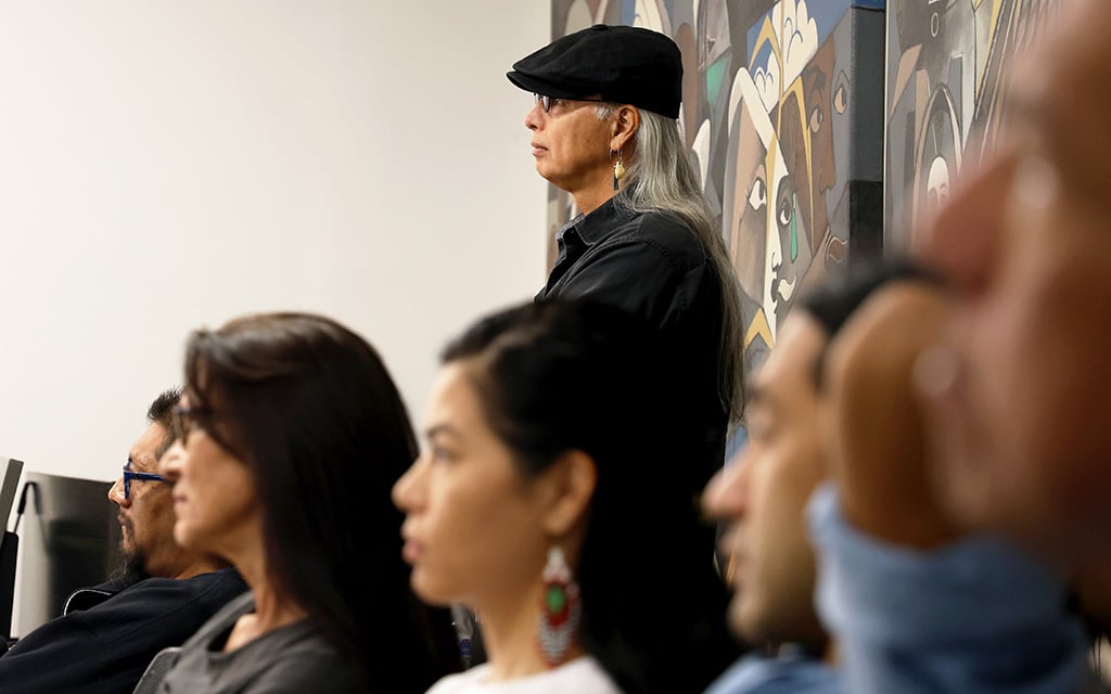 A person with long gray hair stands beside a mural with abstract faces, while others sit in the foreground.