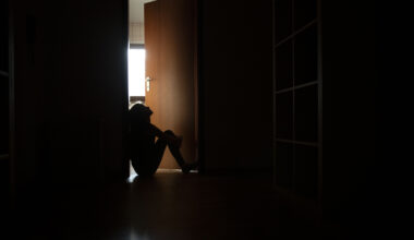 A photo of a teenager sitting in the frame of a doorway backlit by a room filled with daylight.