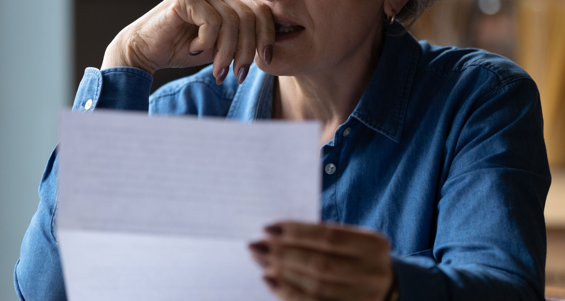 A closely cropped photo of a senior woman holding a paper letter. She presses her hand to her lips as she makes a stressed expression.