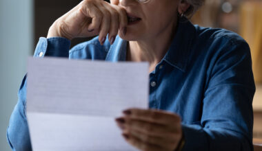 A closely cropped photo of a senior woman holding a paper letter. She presses her hand to her lips as she makes a stressed expression.