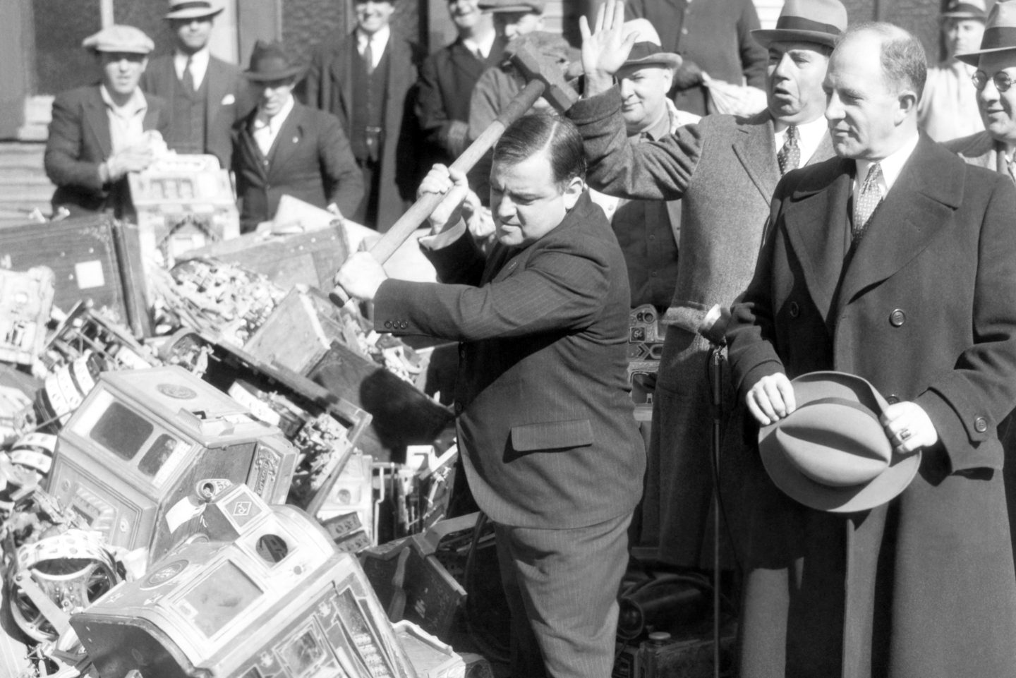 A black and white photo of a man swinging a sledgehammer down on machines with a crowd gathered behind him.