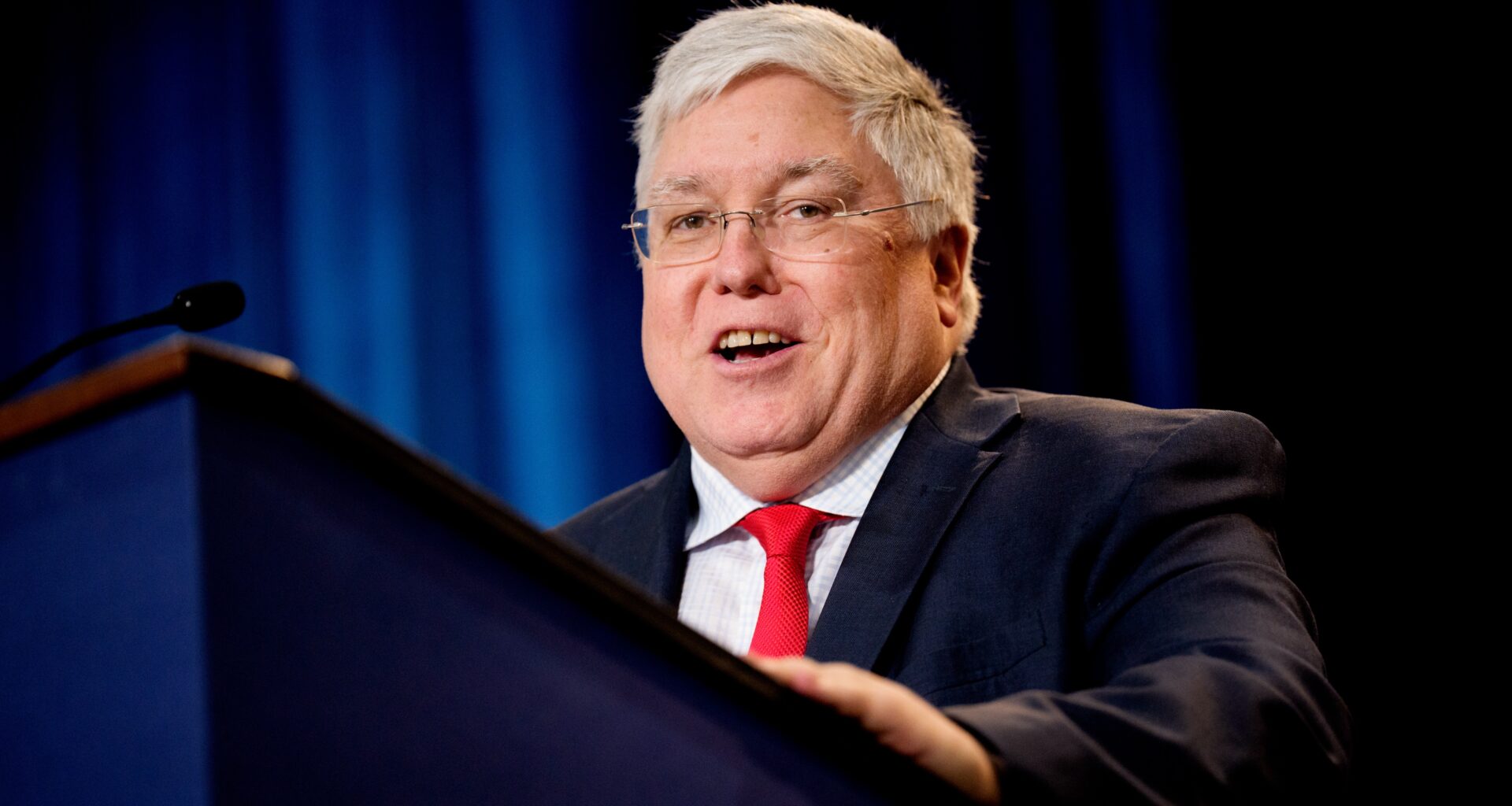 A man with grey hair, glasses, a red tie and a dark suit stands behind a podium