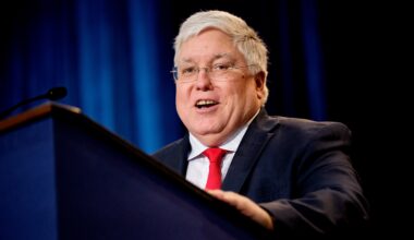 A man with grey hair, glasses, a red tie and a dark suit stands behind a podium