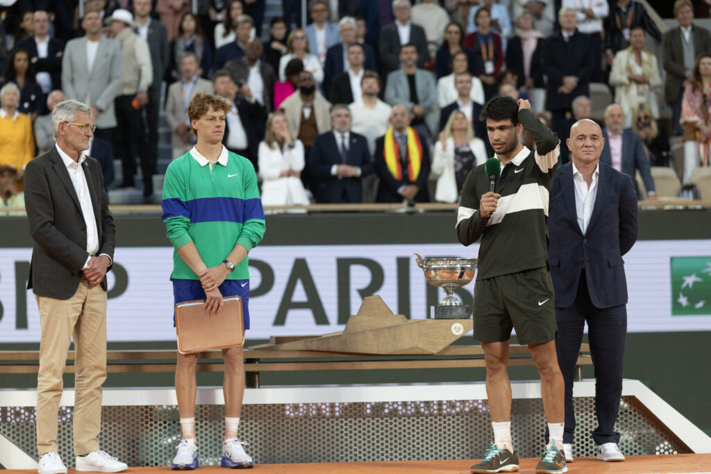 Carlos Alcaraz speaks into a microphone after beating Jannik Sinner, who stands with his runners-up trophy