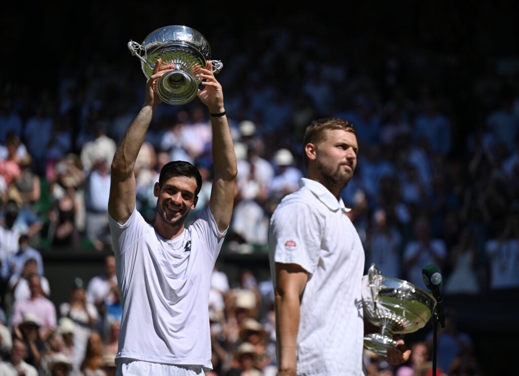 Julian Cash of Great Britain celebrates with his Gentlemen's Doubles Trophy as team mate Lloyd Glasspool of Great Britain looks on following victory against Rinky Hijikata of Australia and David Pel of Netherlands in the Gentlemen's Doubles Final on day thirteen of The Championships Wimbledon 2025 at All England Lawn Tennis and Croquet Club.