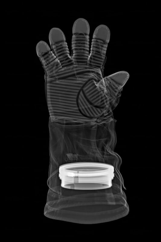 X-ray image of a gloved hand with striped details, holding a round, transparent container at the palm, all set against a black background.