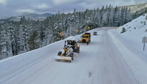 aerial view of two slow plows headed up a snow covered road with snow covered fir trees in the distance