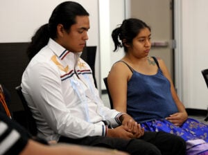 A man and a woman sitting side by side, the man in a white patterned shirt and the woman in a blue top and colorful skirt.